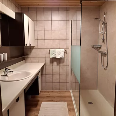 Ein modernes Badezimmer mit einer großen Dusche und einem Waschbecken. Der Raum hat helle Fliesen und eine Holzdecke.