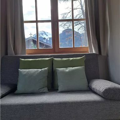 Ein gemütliches Sofa mit grünen Kissen steht neben einem Fenster. Draußen sind Berge und der Himmel ist teilweise bewölkt.