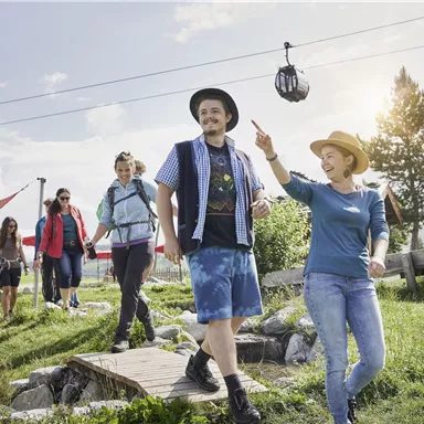 Eine Gruppe von fünf Personen spaziert auf einem Weg in der Natur. Im Hintergrund sind eine Seilbahn und eine sonnige Landschaft zu sehen.