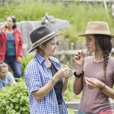 Zwei Frauen stehen in einem Garten und unterhalten sich freundlich. Im Hintergrund sind weitere Personen mit Gemüse beschäftigt.