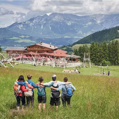Eine Gruppe von fünf Personen steht in einer grünen Wiese und schaut auf ein malerisches Bergpanorama. Im Hintergrund ist ein gemütliches Holzhaus und Spielplätze zu sehen.