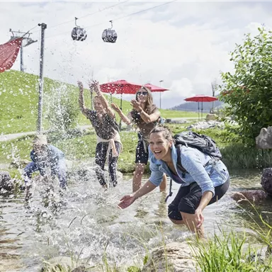 Eine Gruppe junger Menschen spielt im Wasser und spritzt herum. Im Hintergrund sind Gondeln und rote Sonnenschirme sichtbar.