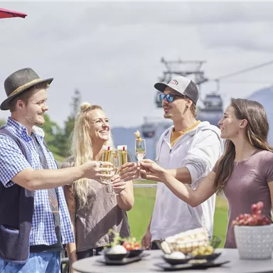 Eine fröhliche Gruppe von vier Personen stößt mit Gläsern an. Im Hintergrund sind Berge und eine Seilbahn zu sehen.