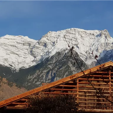 An impressive mountain landscape with snow-capped peaks under a blue sky. In the foreground, a wooden house can be seen.