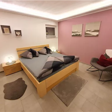 Ein modernes Schlafzimmer mit einem Holzbett und gemütlichen Kissen. Die Wände sind in sanften Farben gehalten, und ein bequemer Sessel steht in der Ecke.