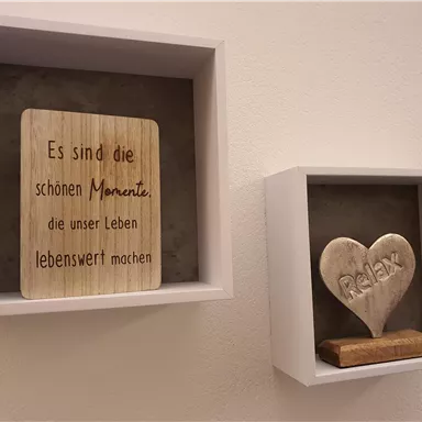Zwei dekorative Rahmen sind an der Wand befestigt. Der linke enthält eine Holztafel mit einem inspirierenden Spruch und der rechte zeigt ein herzförmiges Stück mit dem Wort "Relax".