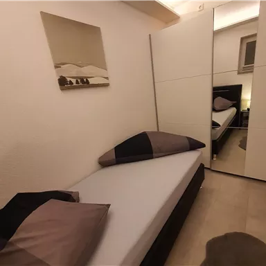 Ein einfaches Schlafzimmer mit einem Einzelbett, großen Kissen und einem Spiegel. An der Wand hängt ein Bild von einer Landschaft.