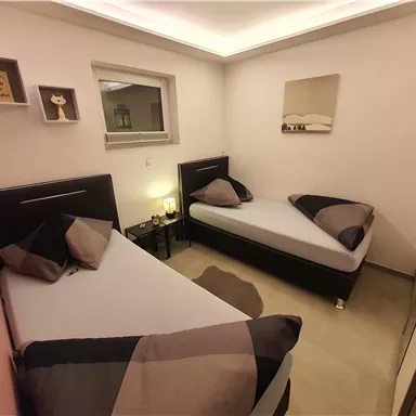 Ein kleines, modernes Zimmer mit zwei Einzelbetten und eleganter Dekoration. Die Wände sind in neutralen Farben gehalten, und es gibt einen Nachttisch mit einer Lampe.