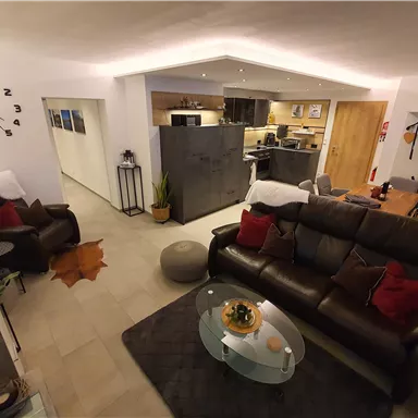 Ein gemütliches Wohnzimmer mit einer großen Ledercouch und dekorativen Kissen. Die offene Küche ist modern und gut ausgestattet, mit einem warmen Ambiente.