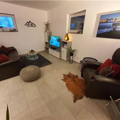 Ein gemütliches Wohnzimmer mit Fernseher, zwei bequemen Sesseln und einer Couch. Die Wände sind mit Bildern dekoriert und es gibt Böden aus hellen Fliesen.