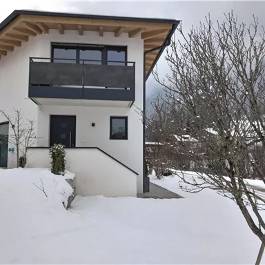 A modern house with a large balcony, surrounded by snow. In the background, bare trees are visible.