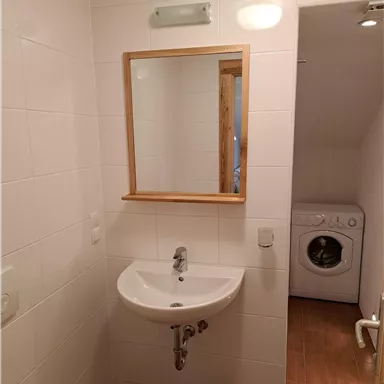 Ein modernes Badezimmer mit einem Waschbecken, einem Spiegel und einer Waschmaschine. Die Wände sind weiß gefliest und es gibt einen Holzfußboden.