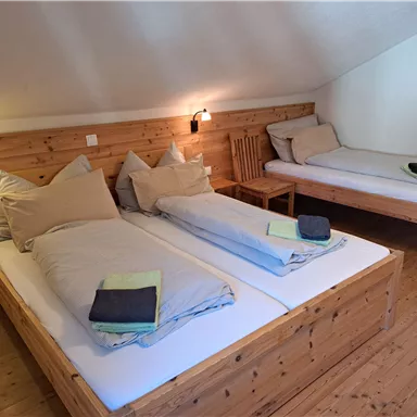 Ein gemütliches Zimmer mit zwei Betten und Holzmöbeln. Die Wände sind hell und die Beleuchtung ist sanft.