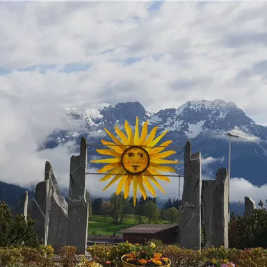 A colorful sun sculpture stands before majestic mountains, partially shrouded in clouds. In the foreground, flowers can be seen that bring the scene to life.