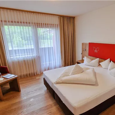 A modern room with a large bed and wooden floor. There is a small table and a red armchair next to the window.