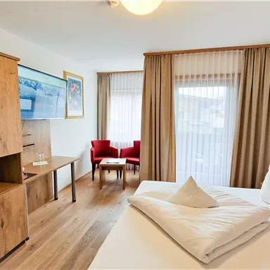 A modern hotel room with a large bed and wooden furniture. There is a small seating area with red chairs and a bright window front.