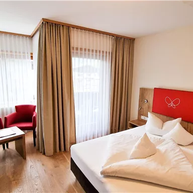 A cozy bedroom with a large bed and red accents. Two chairs and a table stand in front of a window with curtains.