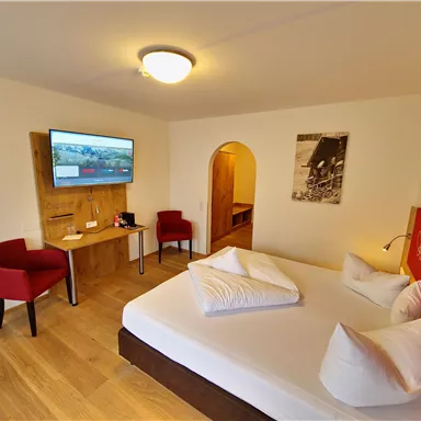 A modern hotel room with a double bed, a television, and a small table. Red chairs and wooden flooring give the room a cozy atmosphere.