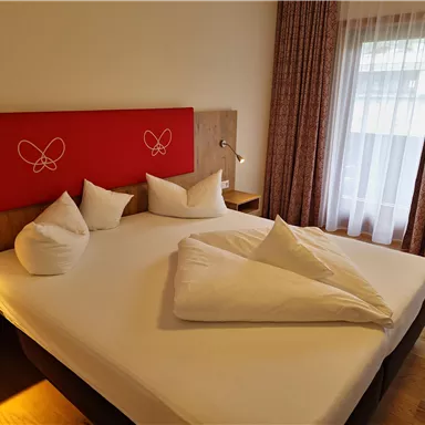 A modern hotel room with a large bed and white pillows. In the background, there are curtains and a window that lets in daylight.