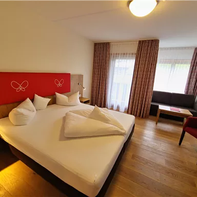 A cozy bedroom with a large bed and several pillows. In the background, a sofa and a table can be seen, as well as large windows with curtains.