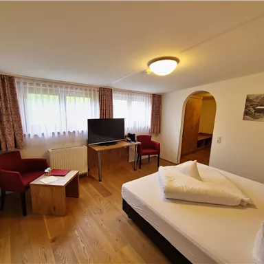 A cozy room with a double bed, a television, and two red chairs. The windows provide plenty of daylight and a welcoming atmosphere.