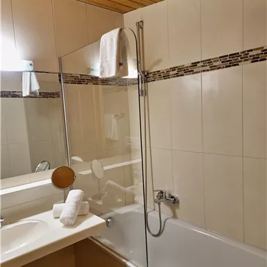A modern bathroom with a bathtub and a glass shower partition. The walls are tiled in light colors, and there are fresh towels on the vanity top.
