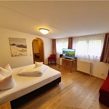 A bright room with a large bed, a television, and a comfortable armchair. Large windows provide plenty of daylight.