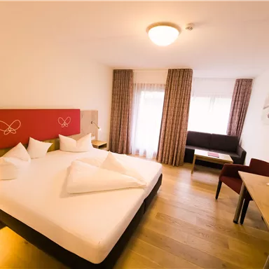 A modern bedroom with a large double bed, wooden floor, and a window that lets in plenty of light. The room features a sofa and a desk.