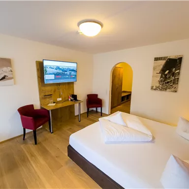 A modern hotel room with a double bed, a television, and two red chairs. Bright walls and wooden floors create an inviting atmosphere.
