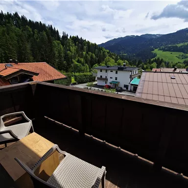 A balcony with two chairs and a table offers a view of green mountains and a small village. The surroundings are surrounded by forests and the sky is partly cloudy.