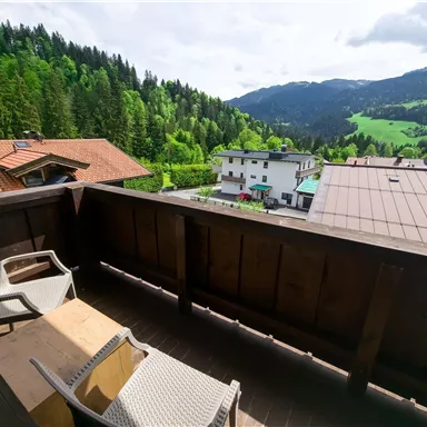 A balcony with two chairs and a table offers a view of green forests and mountains. The atmosphere is calm and inviting.