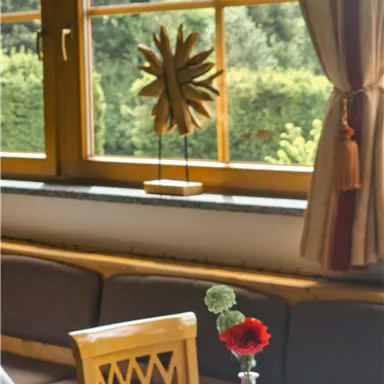 A cozy table with a vase and flowers in a bright room. In the background are windows overlooking green landscapes.