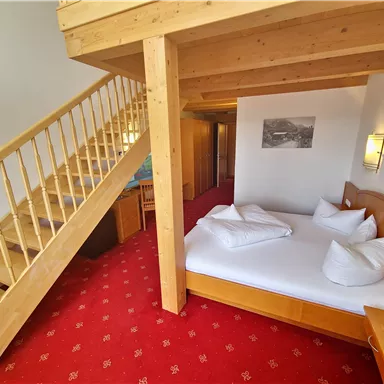 A cozy room with a double bed and a wooden staircase. The floor is red with carpet, and there are wooden decorations on the walls and furniture.
