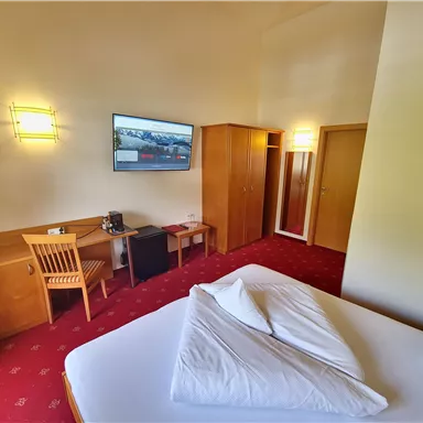 A cozy hotel room with a double bed, a table, and a television. The carpet is red, and there is a wooden wardrobe as well as a chair.