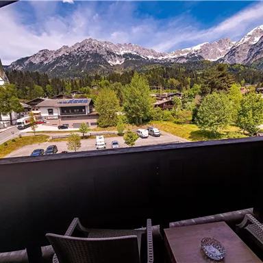 A view from a balcony overlooking the mountains and a small village. In the foreground, chairs and a table can be seen.