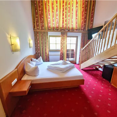 A cozy room with a double bed and red carpet. A staircase leads to an upper area decorated with windows and curtains.