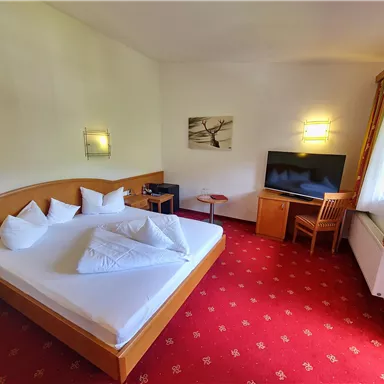 A cozy hotel room with a large bed, a television, and a table. The walls are light and the carpet has a red pattern.