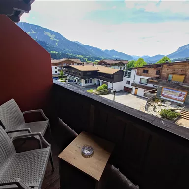 A balcony with two white chairs and a small table. In the background, mountains and houses are visible, with a clear sky.