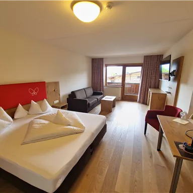 A modern hotel room with a double bed, a couch, and a desk. There are large windows with a view outside and a TV on the wall.