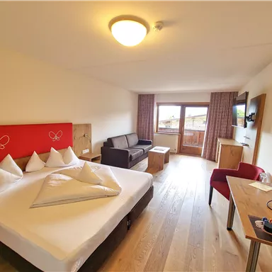 A modern hotel room with a large bed, a sofa, and a table. The room offers access to a balcony with a view.