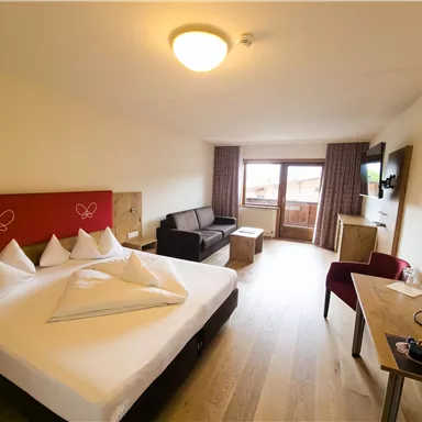 A modern hotel room with a double bed, a seating area, and a balcony. The decor is bright and friendly, with wood-colored floors and comfortable furniture.