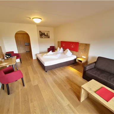 A modern hotel room with a large double bed, a sofa, and a small table. The wooden floors and cozy furnishings create an inviting atmosphere.