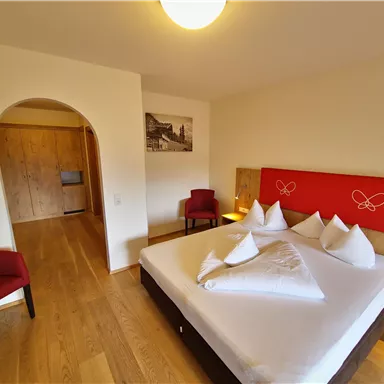 A bright room with a large double bed and red chairs. The walls are light and the floor is made of wood.