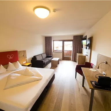 A modern hotel room with two beds, a sofa, and a desk. There are large windows overlooking a balcony.