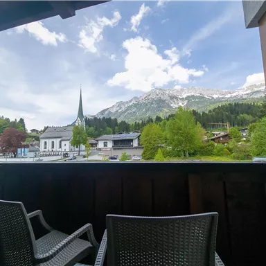 A beautiful view from a balcony over mountains and a small town. In the foreground, two chairs are present, while the landscape in the distance is green and mountainous.
