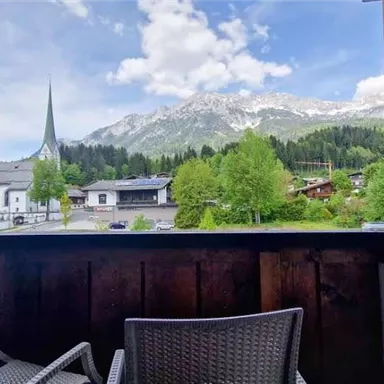 A balcony view of a picturesque mountain landscape with a church and green trees. The sky is partly cloudy, and in the background, majestic mountains are visible.