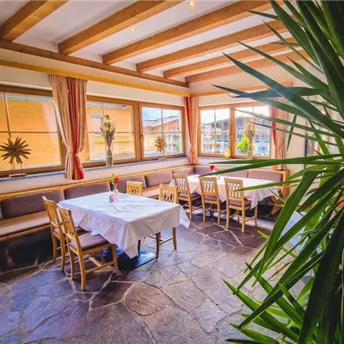 A cozy dining room with wooden ceilings and large windows. The tables are set for guests, and a green plant adds a fresh touch.