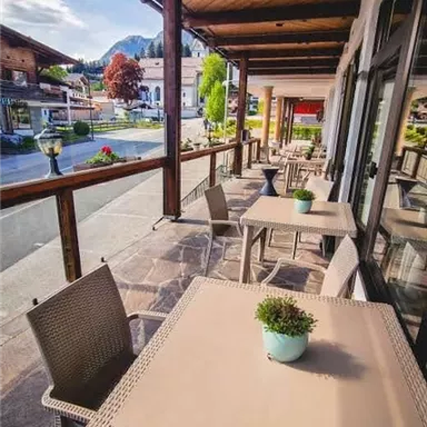 An inviting terrace with several tables and chairs. In the background, green trees and a quiet street can be seen.