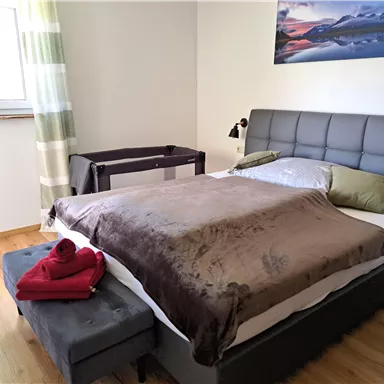 A modern bedroom with a large bed and a cozy duvet. A chair and a table complement the simple, elegant furnishings.