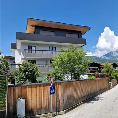 Ein modernes Haus mit einem Balkon und großen Fenstern. Umgeben von grünen Bäumen und einer ruhigen Straße.